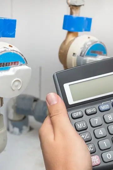 Mão fazendo contas numa calculadora em frente a dois hidrômetros