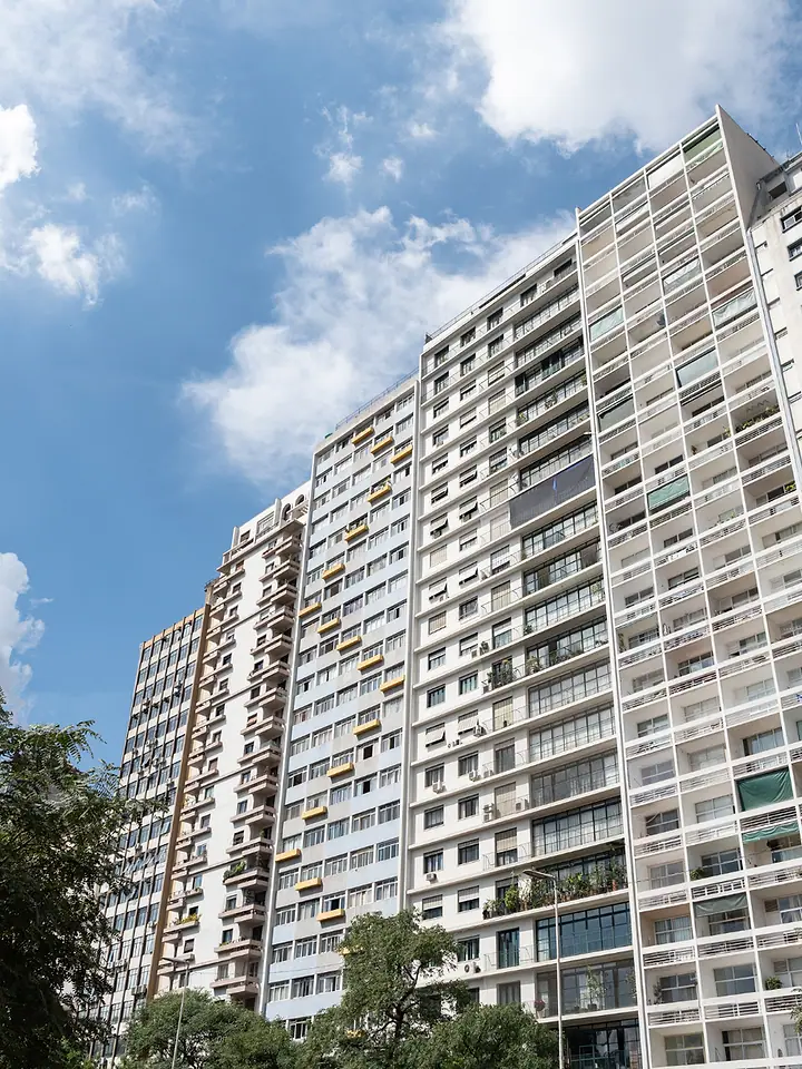 Prédio residencial alto visto de baixo, com céu azul e árvores na frente